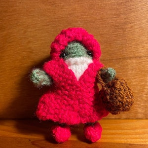 May include: A small crocheted gnome wearing a red hooded robe and carrying a brown bag. The gnome has a green face and white beard.