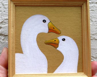 May include: A small square painting in a gold frame featuring two white geese with orange beaks set against a solid gold background. The geese are positioned with one larger goose on the left and a smaller goose on the right, both facing right.