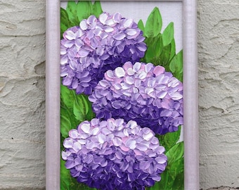 May include: A framed painting featuring three vibrant purple hydrangea blooms with green leaves. The artwork is set against a white background and enclosed in a light-colored wooden frame, creating a visually appealing floral composition.
