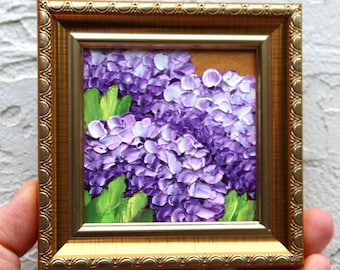 May include: A small square oil painting featuring thick impasto brushstrokes of purple hydrangea flowers and bright green leaves, encased in an ornate metallic gold frame with a decorative scalloped edge and a smooth inner border.