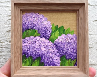May include: A small framed painting of purple hydrangeas with green leaves against a gold background. The artwork is in a natural wood frame. The painting has a textured, impasto style, with visible brushstrokes creating depth and dimension.