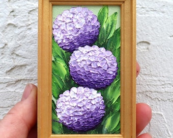 May include: A small rectangular painting in a gold-colored frame featuring three textured purple hydrangea flower heads surrounded by vibrant green leaves. The artwork displays thick impasto brushstrokes and a layered floral composition.