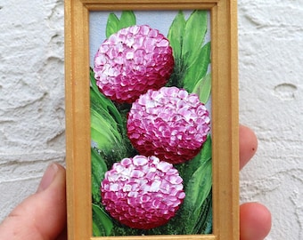 May include: A miniature painting in a rectangular wooden frame. The artwork depicts three vibrant pink flower blooms with textured petals, set against a backdrop of green leaves. The frame has a gold finish, enhancing the artwork's visual appeal.