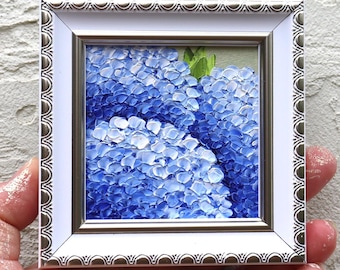 May include: A miniature framed painting of blue hydrangeas. The artwork features a close-up view of the flowers with textured brushstrokes. The frame is white with a silver decorative edge, creating a classic look.