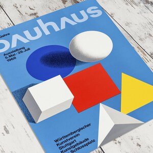 May include: A blue poster with the word "Bauhaus" in white letters. The poster features geometric shapes in red, yellow, white, and blue. The text on the poster reads "Ausstellung 5. Mai - 28. Juli 1968" and "W&uuml;rttembergischer Kunstverein Stuttgart Kunstgeb&auml;ude Schlossplatz."