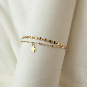 Könnte beinhalten: Zweireihiges goldfarbenes Armband-Set. Die obere Schicht hat eine zarte Kette aus kleinen, flachen, kreisförmigen Gliedern. Die untere Schicht hat eine feine Kette mit einem kleinen Kreuzanhänger. Weißer Hintergrund.