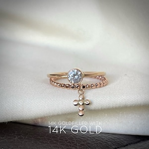 May include: A delicate gold ring set with a single round clear stone, featuring a dangling cross charm. The ring is made of 14k gold filled.