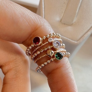 May include: Five gold rings with different colored gemstones. The rings are stacked on a finger and feature a red, white, green, and gold stone.