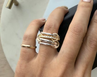 May include: A hand wearing a gold band ring and a multi-layered ring with gold and silver tones. The multi-layered ring features a row of small, clear stones. The rings are displayed against a neutral background.