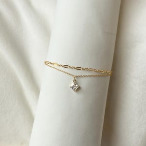 May include: A delicate gold-colored bracelet featuring two fine chains. The top chain has an oval link design, while the lower chain suspends a small, square, clear gemstone pendant. The bracelet is displayed against a white background.