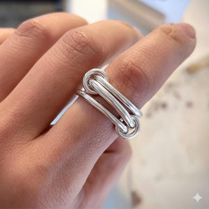 May include: A silver ring with a distinctive linked design. The ring has three parallel bars joined by two circular loops. The polished metal reflects light, creating a modern, minimalist look. The ring is worn on a finger.