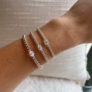 May include: Three gold beaded bracelets with a single clear gemstone accent on each. The bracelets are stacked on a person's wrist.