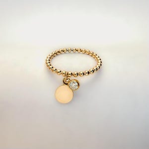 Può includere: Un anello dorato con perline con un piccolo ciondolo rotondo e una singola gemma trasparente. La fascia dell'anello è composta da piccole perline distanziate uniformemente. Il ciondolo è un piccolo cerchio piatto e la gemma è rotonda e incastonata.