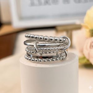 May include: A multi-layered silver ring with various textures, including beaded and smooth bands. The ring has a modern design with a mix of different ring styles, creating a unique and stylish accessory. The ring is displayed on a white cylindrical stand.