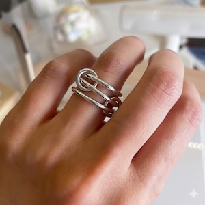 Può includere: Un anello color argento dal design unico. L'anello presenta un elemento superiore circolare collegato a due barre parallele, creando un look moderno e geometrico. L'anello è indossato su un dito, mostrando il suo stile elegante e contemporaneo.