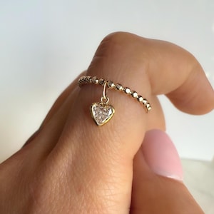 May include: A gold ring with a beaded band and a heart-shaped pendant. The pendant features a clear, faceted stone set within a gold frame. The ring is shown on a finger.
