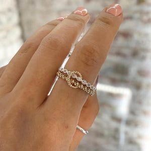 Op de afbeelding: Twee ringen, één goud en één zilver, worden op een vinger getoond. De gouden ring heeft een knoopontwerp en de zilveren ring heeft kleine, heldere stenen. Een andere zilveren ring is zichtbaar op de aangrenzende vinger.