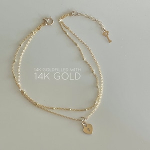 May include: A delicate gold-filled necklace with a double-strand design. The necklace features a heart-shaped lock pendant and a small key charm. The text "14K Goldfilled with 14K Gold" is displayed in the image.