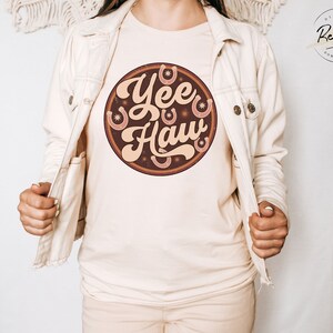 May include: A white t-shirt with a brown circle graphic that says "Yee Haw" in a retro font. The graphic also features several brown horseshoes and stars.