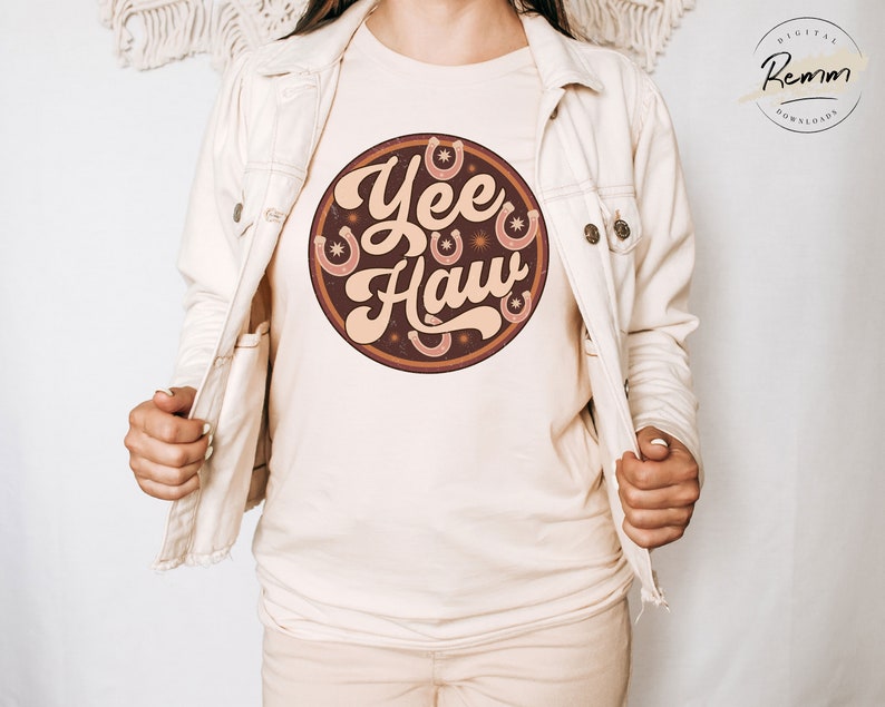 May include: A white t-shirt with a brown circle graphic that says "Yee Haw" in a retro font. The graphic also features several brown horseshoes and stars.