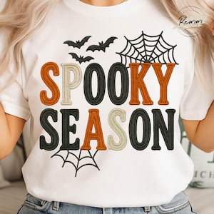 以下が含まれることがあります： オレンジ、黒、クリーム色で「SPOOKY SEASON」と刺繍された白いTシャツ。デザインにはコウモリとクモの巣が含まれており、ハロウィーンに最適です。
