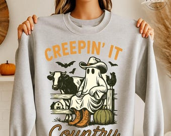 May include: A light gray sweatshirt with the text "CREEPIN' IT COUNTRY" in orange and brown. The graphic features a ghost wearing a cowboy hat and boots, sitting on a fence with a cow, chicken, bats, and a pumpkin.