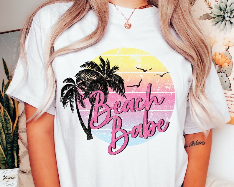 Op de afbeelding: Wit T-shirt met een retro zonsondergang afbeelding met palmbomen en de tekst "Beach Babe" in roze en zwarte letters.