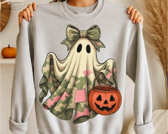May include: A light gray sweatshirt featuring a cartoon ghost in a camouflage pattern with a green bow, holding an orange jack-o'-lantern bucket. The ghost has black eyes and a pink patch. A fun Halloween-themed design.