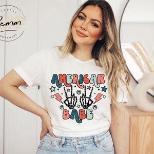 May include: White t-shirt with a colorful graphic design that says "American Babe" with peace signs, lightning bolts, and stars.
