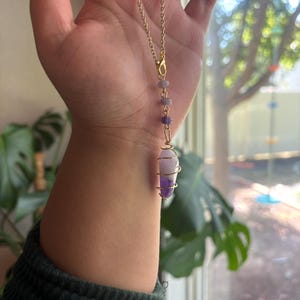 May include: A gold-colored chain necklace with a pendant featuring a purple and white crystal wrapped in gold wire. The necklace also has small purple beads. The background includes a green sweater and a window.