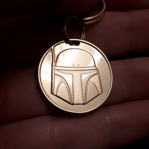 May include: A gold-coloured metal keyring featuring the helmet design of Boba Fett from Star Wars. The round pendant has a detailed, engraved image of the helmet and is attached to a gold-coloured ring.