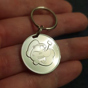 May include: Silver keychain featuring a cartoon image of Popeye the Sailor Man with his signature pipe and a wink.