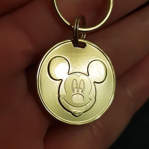 May include: A gold-coloured metal keychain featuring a Mickey Mouse head design. The keychain is round and has a smooth, shiny surface.