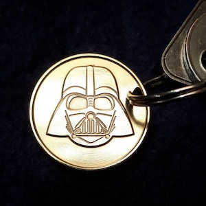 Puede incluir: Un llavero dorado con una imagen redonda grabada del casco de Darth Vader.