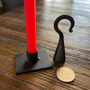 May include: A black metal candle holder with a red candle and a black metal hook. The candle holder is square and has a small base. The hook is curved and has a pointed end. A US quarter is included for scale.