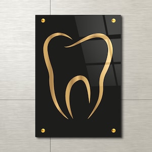 Puede incluir: Un contorno de diente dorado sobre un fondo negro. El diente es un contorno simple y está centrado en el fondo negro. El fondo es una superficie de acrílico negro brillante.