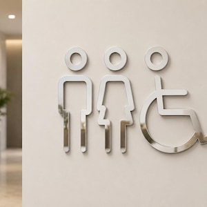 Pode incluir: Sinais de banheiro prateados para homens, mulheres e pessoas em cadeiras de rodas. Os sinais são montados em uma parede bege clara. Os símbolos são minimalistas e modernos, com um acabamento metálico reflexivo. Os sinais são fáceis de reconhecer.