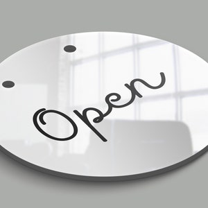 Op de afbeelding: Een wit rond bord met het woord "Open" in zwarte cursieve letters. Het bord heeft twee kleine zwarte stippen bovenaan.