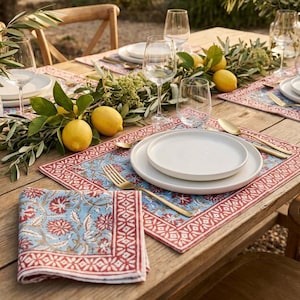 Puede incluir: Una mesa de madera rústica preparada para comer al aire libre. La mesa está adornada con platos blancos, cubiertos dorados y manteles individuales y servilletas estampadas en azul y rojo. Limones frescos y vegetación añaden un toque natural, con copas de vino y un jarrón de flores.