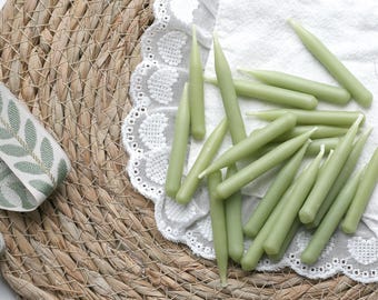 May include: A collection of pale green taper candles arranged on a white lace-edged doily. The candles are scattered across the doily, with a woven placemat and a ribbon with a leaf pattern in the background. The candles are approximately 6 inches long.