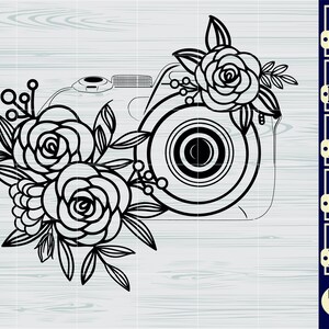 May include: Black and white line art illustration of a camera with floral embellishments. The camera has a lens and a body with a floral design on the side. The flowers are stylized roses with leaves and stems.