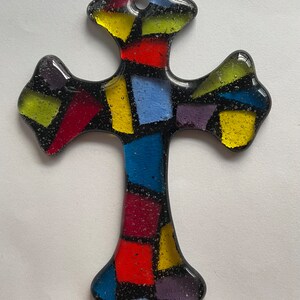 May include: A stained glass cross with a mosaic design. The cross is made of small pieces of glass in various colors, including red, yellow, blue, and purple. The glass is set in a black frame.