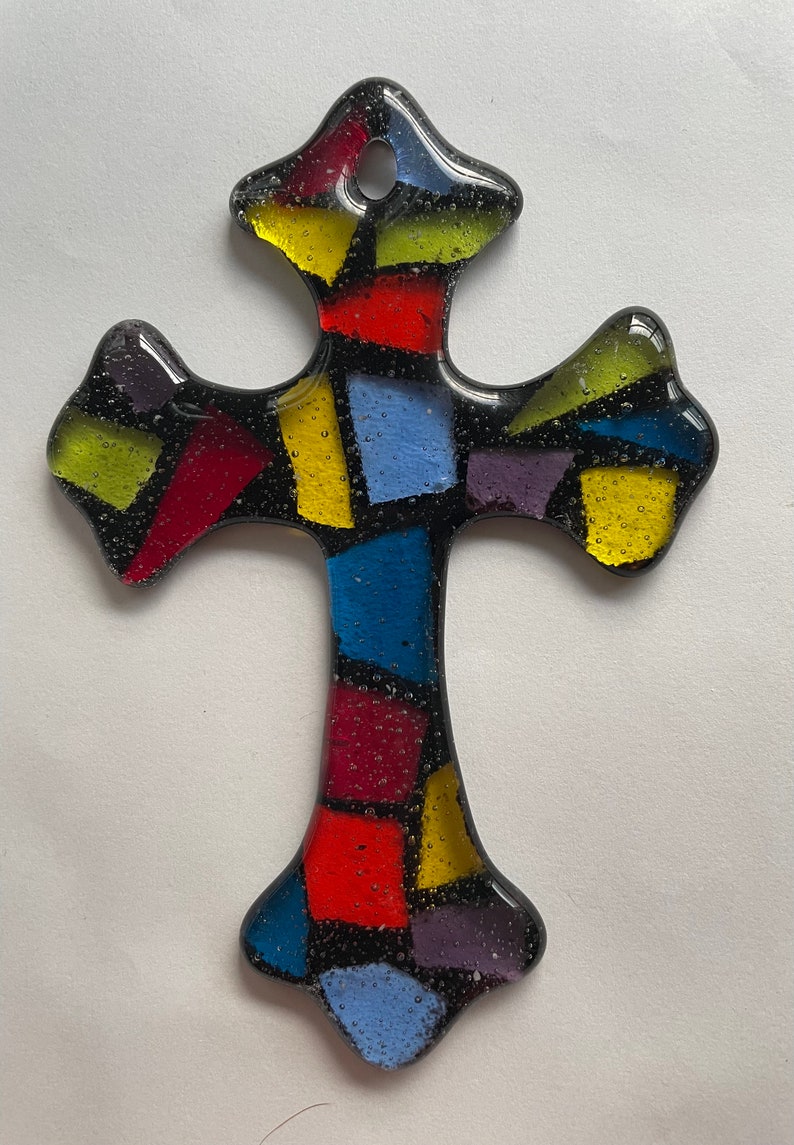 May include: A stained glass cross with a mosaic design. The cross is made of small pieces of glass in various colors, including red, yellow, blue, and purple. The glass is set in a black frame.