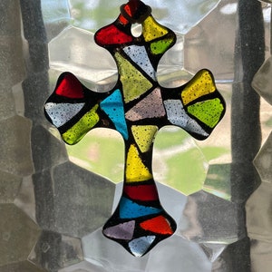 May include: A colorful stained glass cross with a mosaic design in shades of red, yellow, green, blue, and purple. The cross is hanging from a white string.