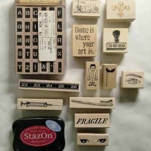 May include: A collection of wooden stamps and a black ink pad. Stamps include letters, phrases like "Home is where your art is," and various illustrations. The ink pad is labeled "StazOn Blazing Red."