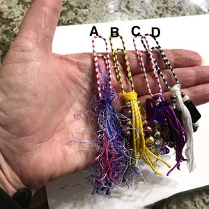 May include: Five colorful paper clips with decorative tassels and beads. The paper clips are red and white striped, black and white striped, and yellow and black striped. The tassels are purple, yellow, white, and black. The beads are purple, gold, and silver.