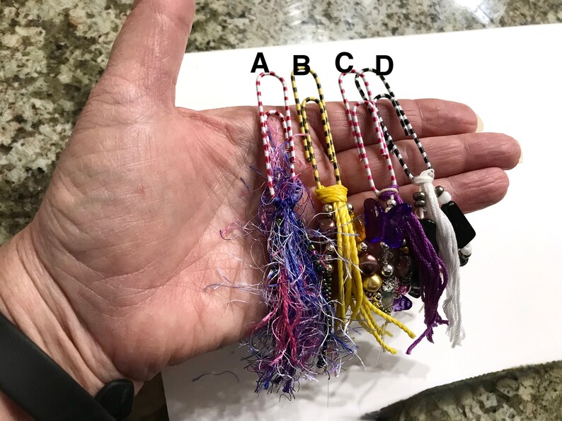 May include: Five colorful paper clips with decorative tassels and beads. The paper clips are red and white striped, black and white striped, and yellow and black striped. The tassels are purple, yellow, white, and black. The beads are purple, gold, and silver.
