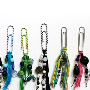 May include: A collection of 12 colorful paperclip charms with various beads, tassels, and embellishments. Each charm features a silver paperclip with a unique combination of colors and textures.