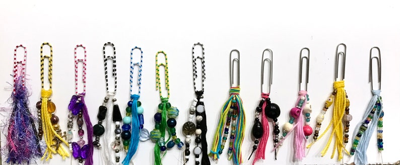 May include: A collection of 12 colorful paperclip charms with various beads, tassels, and embellishments. Each charm features a silver paperclip with a unique combination of colors and textures.