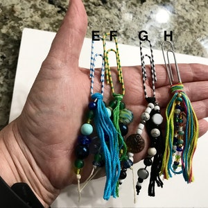 May include: Four colorful beaded and tassel bookmarks made with paper clips. Each bookmark has a different color scheme and bead combination. The bookmarks are held in a hand.